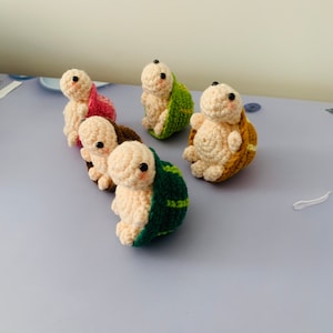 Pode incluir: Cinco brinquedos de tartaruga de crochê em diferentes cores. As tartarugas estão todas em fila, olhando para a câmera. As tartarugas são feitas de fio e têm pequenos olhos pretos.
