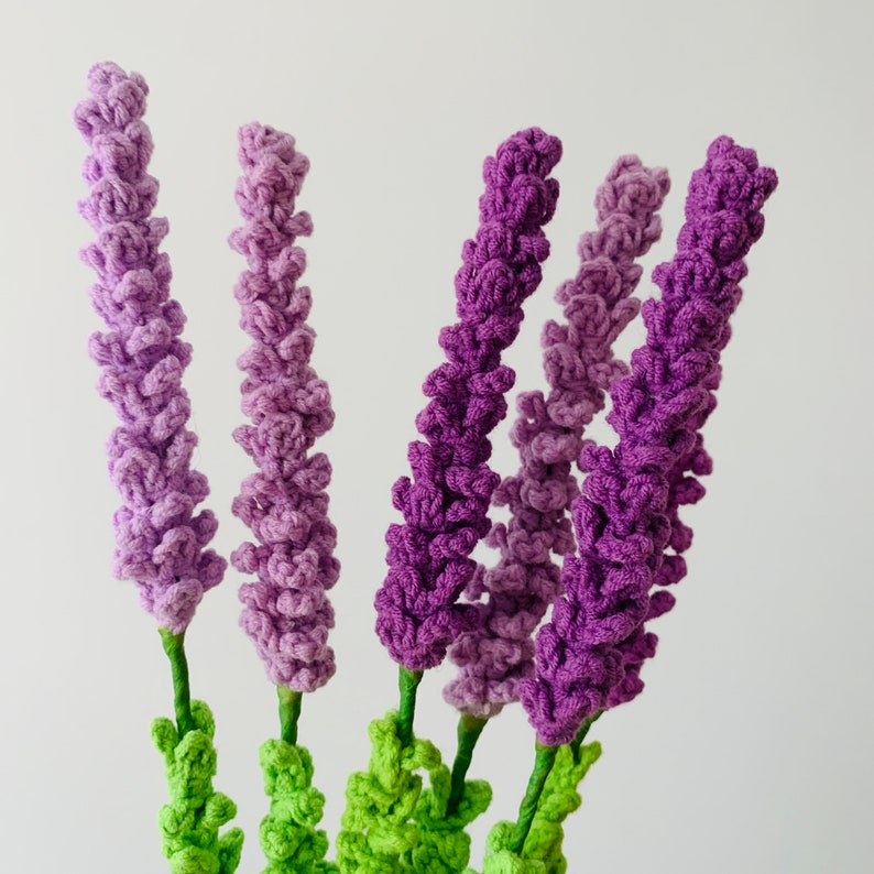 May include: A bouquet of crocheted lavender flowers in shades of purple and green. The flowers are arranged in a vase and are ready to be displayed in a home or office.