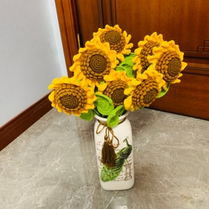 May include: A bouquet of crocheted sunflowers in yellow and brown tones arranged in a white ceramic vase with a peacock design. The vase has the text "La vie est belle" printed on it.