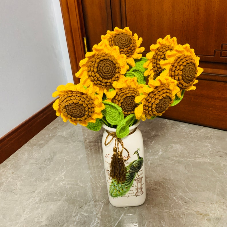 May include: A bouquet of crocheted sunflowers in yellow and brown tones arranged in a white ceramic vase with a peacock design. The vase has the text "La vie est belle" printed on it.