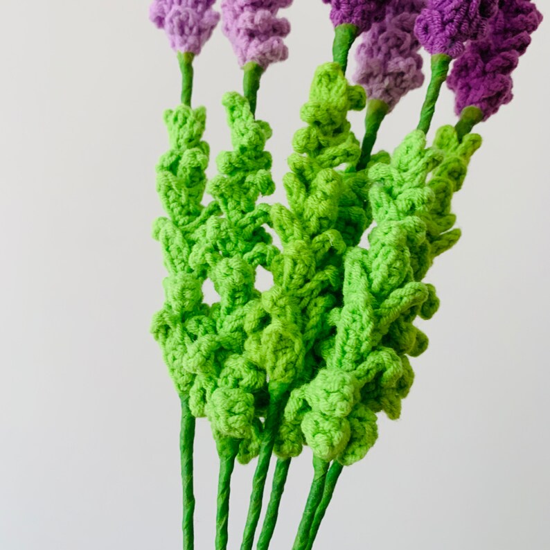 May include: A bunch of crocheted lavender flowers with green stems. The flowers are made with a textured yarn and have a realistic look.
