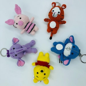 May include: Five handmade crochet keychains featuring cartoon characters. The keychains include a pink pig with black stripes, an orange tiger, a purple elephant, a yellow bear with a red scarf, and a blue creature.