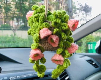 Op de afbeelding: Een gehaakte hangplant met groene bladeren en roze bloemen. De plant zit in een bruine pot en hangt aan een touwtje.