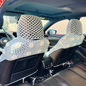 Op de afbeelding: Twee gehaakte autostoelhoezen met een lichtblauwe achtergrond en witte margrietaccenten. De hoezen zijn gemaakt van een delicate, opengeweven stof en hebben een eenvoudig, elegant ontwerp.
