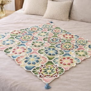 May include: A colourful, handmade crochet blanket with a floral design. The blanket has a repeating pattern of flowers in pink, blue, green, and yellow, with a white border. The blanket is displayed on a bed with neutral-coloured bedding.