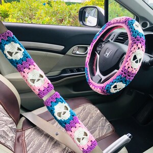 May include: A pink, purple, and blue crocheted steering wheel cover with a skull design. A matching crocheted seatbelt cover with a skull design is also visible.