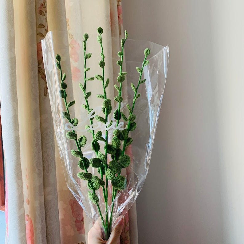 K&ouml;nnte beinhalten: Ein Strau&szlig; gr&uuml;ner geh&auml;kelter Blumen, verpackt in durchsichtigem Plastik. Die Blumen sind in einer Vase arrangiert und haben ein nat&uuml;rliches, organisches Aussehen.
