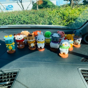 May include: A collection of 12 crocheted animal figurines, each with a different design and colour. The figurines are displayed on a car dashboard, with each one sitting on a small, round base.