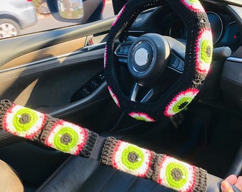 May include: A black crocheted steering wheel cover with white and green eye designs. A matching black, white, and green crocheted seatbelt cover with eye designs.