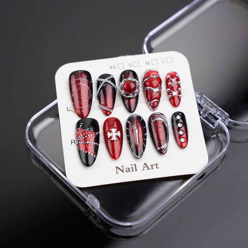 May include: A set of ten artificial nails in a clear plastic case. The nails are mainly red and black, with various designs including silver accents, gems, and a cross. The card reads "Nail Art".