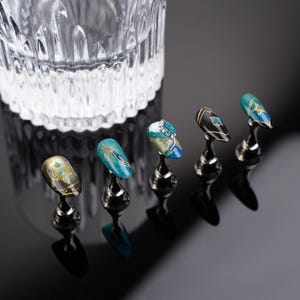 May include: Five decorative nail tips on display stands. The tips are painted with intricate designs featuring gold, blue, and green colors. The tips are arranged in a row on a black surface.