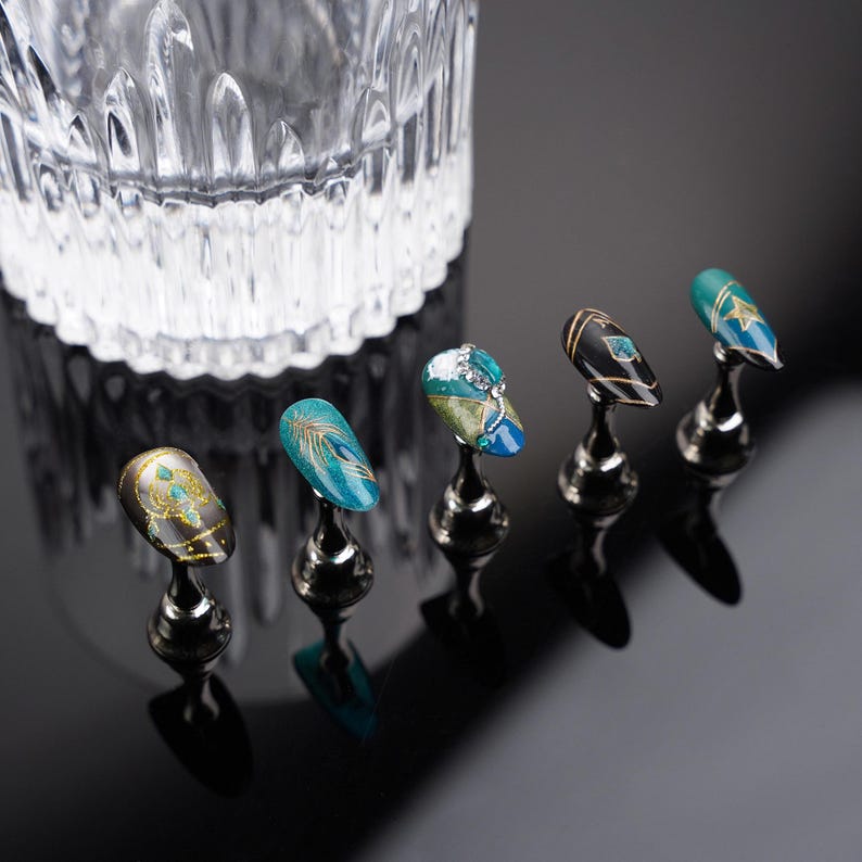 May include: Five decorative nail tips on display stands. The tips are painted with intricate designs featuring gold, blue, and green colors. The tips are arranged in a row on a black surface.