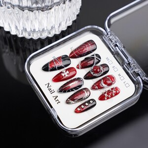 May include: A selection of press-on nail art presented in a clear plastic case. The nails showcase diverse designs in red, black, and silver, incorporating patterns, gems, and chains. The text "Nail Art" is printed on the packaging.