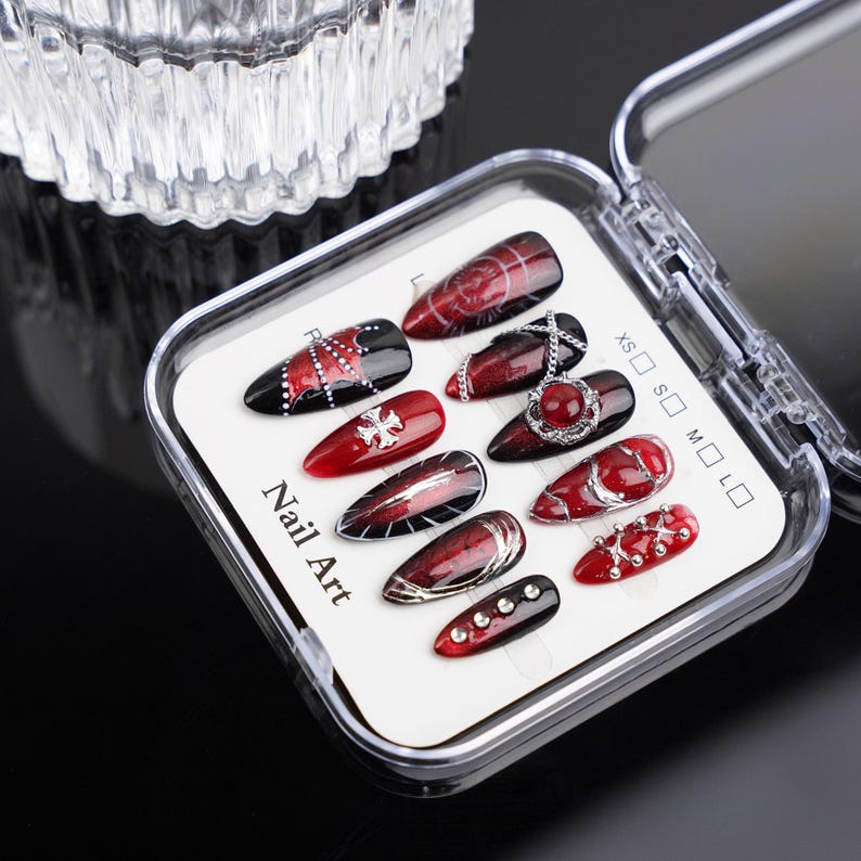 May include: A selection of press-on nail art presented in a clear plastic case. The nails showcase diverse designs in red, black, and silver, incorporating patterns, gems, and chains. The text "Nail Art" is printed on the packaging.