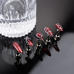 May include: Five artificial nails with elaborate designs, presented on small silver stands. The nails are a deep red hue, adorned with silver chains, gems, and patterns. A clear glass container is visible in the background.