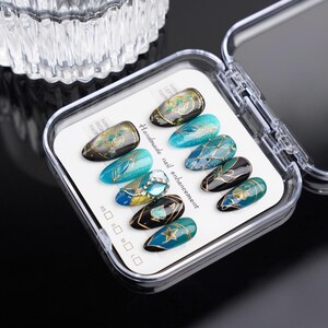 May include: A set of ten false nails with intricate designs featuring blue, green, gold, and black colors. The nails are arranged in a clear plastic case with a white card that reads "Handmade nail enhancement".