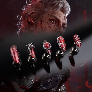 May include: Five red and black nail designs, each displayed on a silver stand. The nails are decorated with silver details and a central red gem. The background shows a person with white hair and red accents.