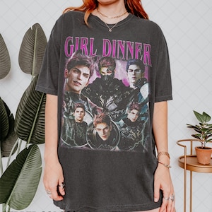 Può includere: Una t-shirt a maniche corte grigio antracite con la scritta GIRL DINNER in lettere rosa brillante sopra un collage grafico di cinque personaggi maschili in abiti scuri. La maglietta ha un girocollo e una vestibilità ampia e rilassata.