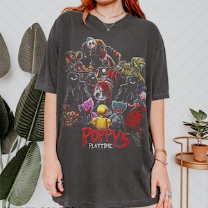 Puede incluir: Camiseta gris oscuro con un gráfico colorido de personajes de Poppy Playtime 5. El diseño presenta varias figuras monstruosas en tonos rojos, morados, amarillos y verdes. El texto "POPPY 5 PLAYTIME" está impreso en rojo.