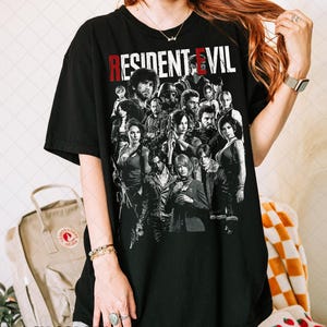 May include: Black t-shirt featuring a group of characters from the video game "Resident Evil" in a grayscale graphic. The title "RESIDENT EVIL" is printed across the top in red and white letters. The words "BLACK COLORS" are printed at the bottom.