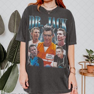 May include: Charcoal grey t-shirt with a vintage-style graphic of a man, and the text "DR. JACK Abbott". The graphic includes multiple images of the man in different poses, wearing an orange vest with "PRIMARY EMERGENCY MD" printed on it.