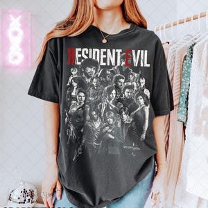 May include: A dark gray t-shirt featuring a group of characters from the Resident Evil video game series. The text "RESIDENT EVIL" is printed across the top in white and red. The words "GRAPHITE COLORS" are printed at the bottom.