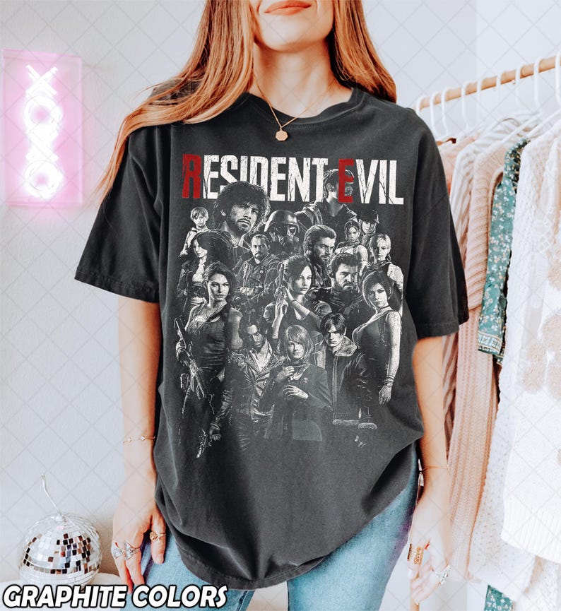 May include: A dark gray t-shirt featuring a group of characters from the Resident Evil video game series. The text "RESIDENT EVIL" is printed across the top in white and red. The words "GRAPHITE COLORS" are printed at the bottom.