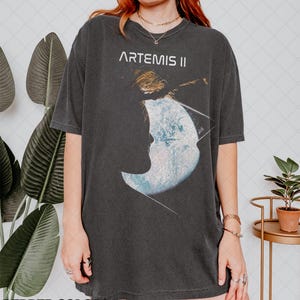 Puede incluir: Camiseta gris oscuro con el texto "ARTEMIS II" sobre un gráfico de la luna y una nave espacial. La camiseta tiene un aspecto vintage. Las palabras "PEPPER COLORS" están en la parte inferior de la imagen.