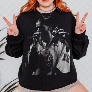 May include: Dark grey sweatshirt with a black and white image of a person holding a microphone, with a signature and the number 18. The person is wearing a jersey with the word "ONTO" visible.