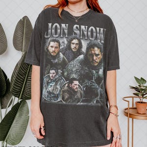 Può includere: Una maglietta nera con una grafica in stile vintage di Jon Snow e altri personaggi della serie televisiva Game of Thrones. Il testo "JON SNOW" è stampato in lettere bianche sulla maglietta.