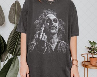 Puede incluir: Una camiseta de manga corta gris carbón con un estampado gráfico blanco de un personaje despeinado con cabello alborotado y maquillaje oscuro en los ojos, haciendo un gesto con el dedo medio. La camiseta tiene un corte relajado y cuello redondo.