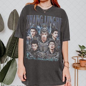 May include: Dark gray t-shirt featuring a graphic of five men in ornate costumes with the text "ZHANG LINGHE" in blue. The shirt is a vintage style with a crew neck and short sleeves.