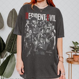 May include: Dark gray t-shirt featuring a group of characters from the Resident Evil video game series. The text "RESIDENT EVIL" is printed across the top in red and white. The shirt is from Pepper Colors.