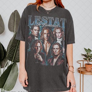 May include: Dark gray t-shirt featuring a graphic of multiple people with the text "LESTAT De Lioncourt" in blue. The shirt has a vintage, distressed look, with the words "PEPPER COLORS" at the bottom.