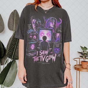 May include: A charcoal gray short-sleeve t-shirt featuring a graphic print for the film I Saw the TV Glow. The design includes two faces, a person sitting before a glowing television, and text that reads There's still time, Reality is a trap.