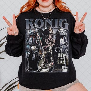 May include: Black sweatshirt with the word "KONIG" in a distressed, silver font. The graphic features a military-themed design with multiple figures in tactical gear, including helmets and weapons. The overall aesthetic is vintage-inspired.