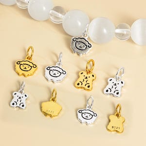 Puede incluir: Una colección de pequeños colgantes decorativos en tonos dorados y plateados. Los colgantes presentan diseños de ovejas y osos, con un lazo para sujetarlos a las joyas. Algunos colgantes tienen la marca "S925".