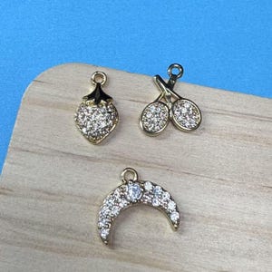 May include: Three gold-tone charms with clear rhinestone accents. The charms are shaped like a strawberry, a pair of tennis rackets, and a crescent moon.