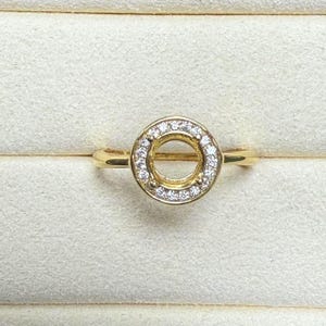 Könnte beinhalten: Eine goldene Ringfassung mit einer runden Öffnung für einen zentralen Stein und einem Heiligenschein aus kleinen, klaren Steinen.