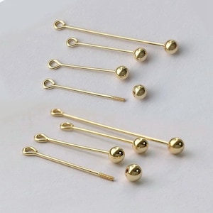 Peut inclure: Ensemble de fournitures de bijoux dor&eacute;es, comprenant des &eacute;pingles droites avec des extr&eacute;mit&eacute;s en boucle et des accents en forme de boule. Plusieurs &eacute;pingles ont des extr&eacute;mit&eacute;s filet&eacute;es. Les fournitures sont dispos&eacute;es sur un fond clair.