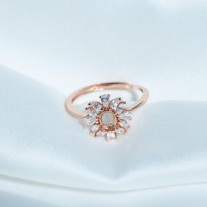 May include: Rose gold ring with a floral design. The ring features a central oval setting surrounded by numerous clear, sparkling stones. The band is a simple, polished rose gold.