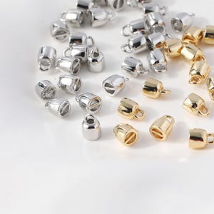 Könnte beinhalten: Eine Ansammlung kleiner, glockenförmiger Kordelenden aus Metall in Silber- und Goldtönen. Jedes Stück verfügt über eine glatte, abgerundete Kappe mit einer kleinen Öse an der Oberseite zur Befestigung, konzipiert für Schmuckkordeln oder Bastelarbeiten.
