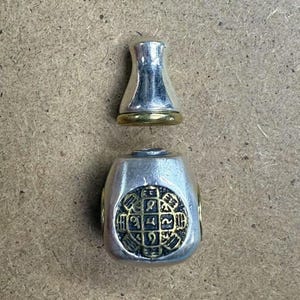May include: A silver and gold metal bottle stopper with a decorative design. The stopper has a round top and a square base. The design on the stopper is a circle with numbers and symbols.