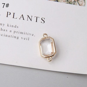 May include: A clear, rectangular crystal pendant with a gold-coloured frame. The pendant has a loop at the top and bottom for attaching to a chain or other jewellery components. The word "PLANTS" is visible in the background.