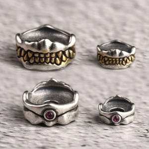 May include: Four silver-toned rings with unique designs. Two rings feature a gold-colored toothy grin, while the other two have a small, round, pink gemstone. The rings are displayed on a textured, light gray surface.
