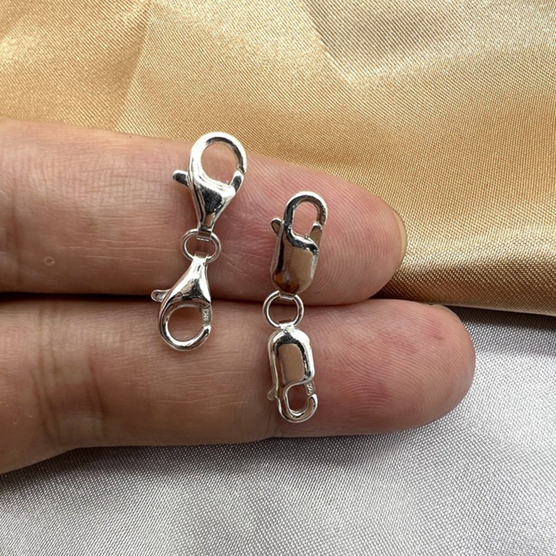 Puede incluir: Dos cierres de langosta de plata, uno con un pivote, que se muestran en una mano. Los cierres se utilizan para la fabricaci&oacute;n de joyas.