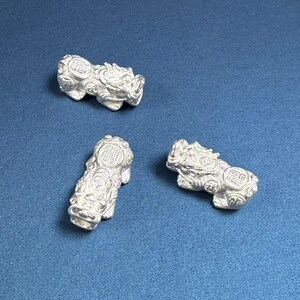 Op de afbeelding: Drie zilverkleurige beeldjes van mythische wezens, mogelijk leeuwen of draken, met ingewikkelde details en een getextureerd oppervlak. De beeldjes zijn gerangschikt op een blauwe achtergrond.