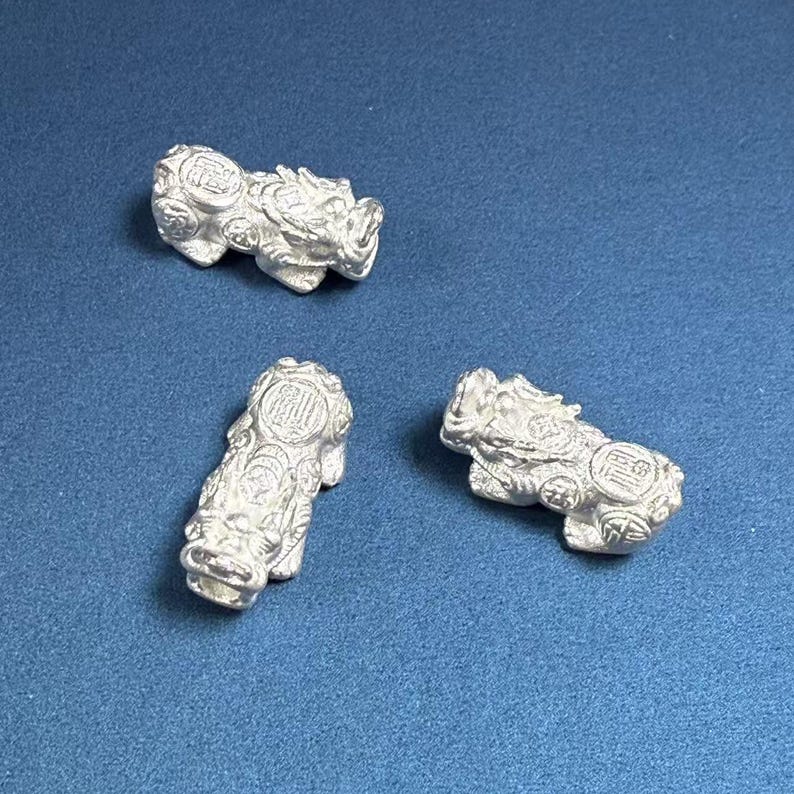Op de afbeelding: Drie zilverkleurige beeldjes van mythische wezens, mogelijk leeuwen of draken, met ingewikkelde details en een getextureerd oppervlak. De beeldjes zijn gerangschikt op een blauwe achtergrond.