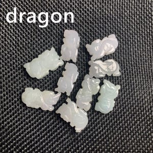 Pode incluir: Uma cole&ccedil;&atilde;o de pequenas figuras de drag&atilde;o de jade esculpido em tons de verde p&aacute;lido e lavanda. A palavra "drag&atilde;o" est&aacute; impressa em branco no topo. As figuras est&atilde;o dispostas sobre uma superf&iacute;cie escura e texturizada.