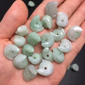 Pode incluir: Uma cole&ccedil;&atilde;o de pequenos pingentes de jade esculpidos em v&aacute;rias formas, incluindo p&aacute;ssaros e formas abstratas. Os pingentes s&atilde;o principalmente verde-claro e brancos, com um acabamento liso e polido. Os pingentes s&atilde;o segurados em uma m&atilde;o.